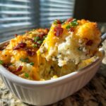 A spoonful of cheesy, bacon-topped mashed potato casserole being lifted from a white baking dish.