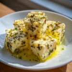 Close-up of cubes of marinated cheese appetizer soaking in olive oil with visible green herbs and spices.