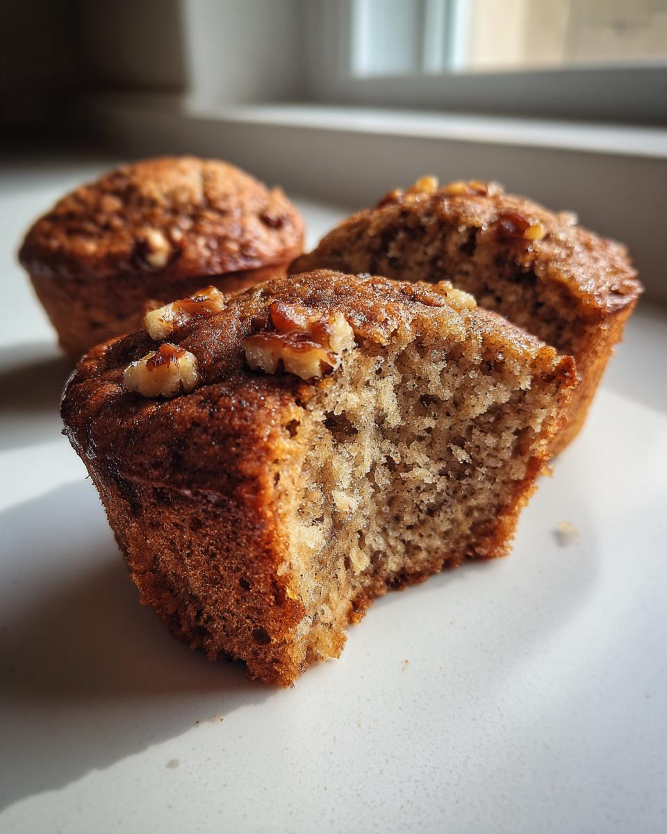 Close-up of a moist banana nut muffin with a bite taken out, showing the crumb texture and walnuts on top.