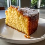 A close-up, sunlit slice of moist olive oil cake dusted with powdered sugar on a white plate.