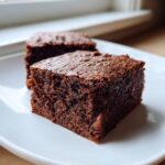 Two rich, dark squares of moist sweet potato brownies served on a bright white plate near a window.