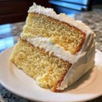 A close-up of a moist slice of yellow vanilla cake recipe layered with white frosting on a white plate.