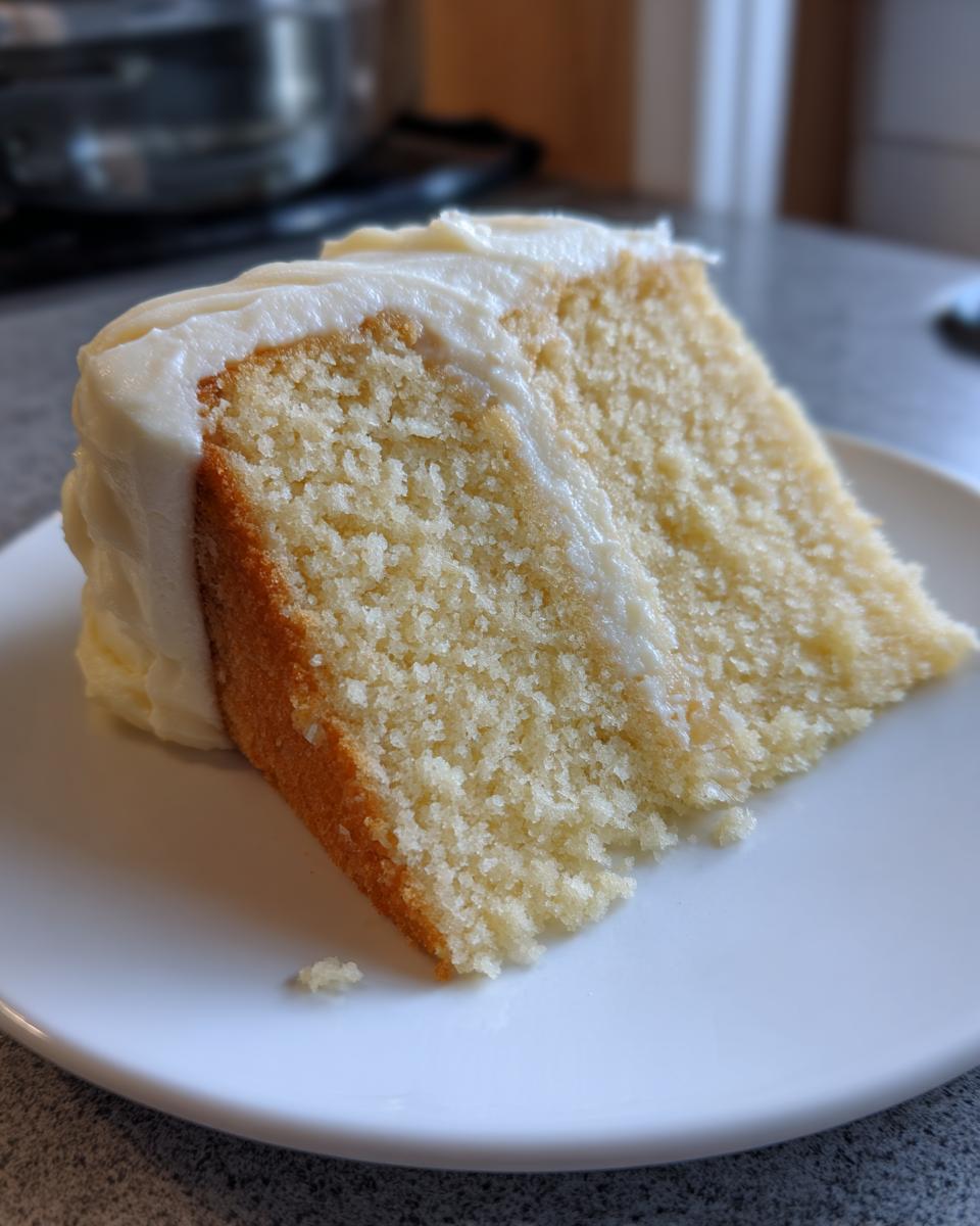 Close-up of a thick, moist slice of vanilla cake with white frosting, showcasing the tender crumb.