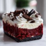 A rich slice of red velvet cheesecake featuring a dark crust, bright red layer, white cheesecake layer, and whipped cream topping with chocolate shavings.