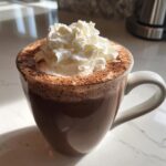 Close-up of a rich, dark hot chocolate topped with a generous swirl of whipped cream and cocoa powder dusting.
