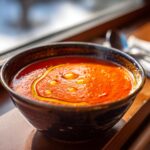 A close-up of rich, vibrant roasted tomato soup in a dark bowl, topped with droplets of golden olive oil.