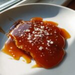 Close-up of rich, glossy caramel sauce topped with flaky sea salt, illuminated by sunlight.