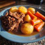 A plate of tender, shredded slow cooker pot roast served with whole cooked potatoes and bright orange carrots in rich gravy.