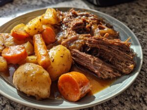 A plate of tender, shredded slow cooker pot roast served with whole potatoes and carrots in rich gravy.