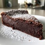 A decadent slice of flourless chocolate torte, dense and dark, dusted generously with powdered sugar on a white plate.