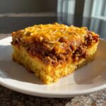 A square slice of hearty tamale pie featuring a thick cornbread base topped with seasoned ground beef and melted cheddar cheese.