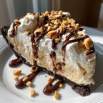 A generous slice of peanut butter ice cream pie with a chocolate cookie crust, topped with whipped cream, chocolate syrup, and chopped peanuts.