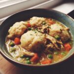 Close-up of a bowl of rich slow cooker chicken and dumplings with visible shredded chicken, carrots, and peas.