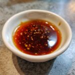 Close-up of a small white bowl filled with dark, glossy Dumpling Sauce featuring visible chili flakes and sesame seeds.