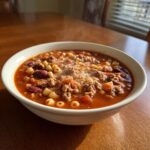 A steaming white bowl filled with rich, tomato-based pasta fagioli soup containing ground meat, kidney beans, and ditalini pasta.