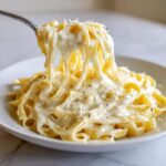 A fork lifting fettuccine noodles coated in rich, creamy homemade alfredo sauce, topped with Parmesan.