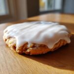 A single, round sugar cookie generously topped with thick, glossy white sugar cookie icing, sitting on a wooden surface.