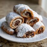 A stack of freshly baked Rugelach pastries dusted heavily with powdered sugar, showing the rich, nutty filling inside.
