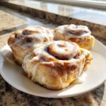 Three freshly baked cinnamon buns generously topped with white icing, sitting on a white plate.