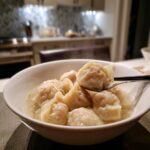A close-up of hot Traditional Chinese Dumplings served in a white bowl of clear broth, with one dumpling lifted by chopsticks.