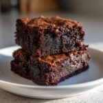Close-up of two intensely dark and moist fudgy brownies stacked on a small white plate.