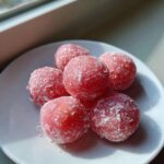 A small pile of bright pink, round watermelon mochi dusted heavily with white powdered sugar on a white plate.