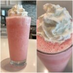 A tall glass filled with a frosty, pink lemonade drink topped generously with whipped cream.