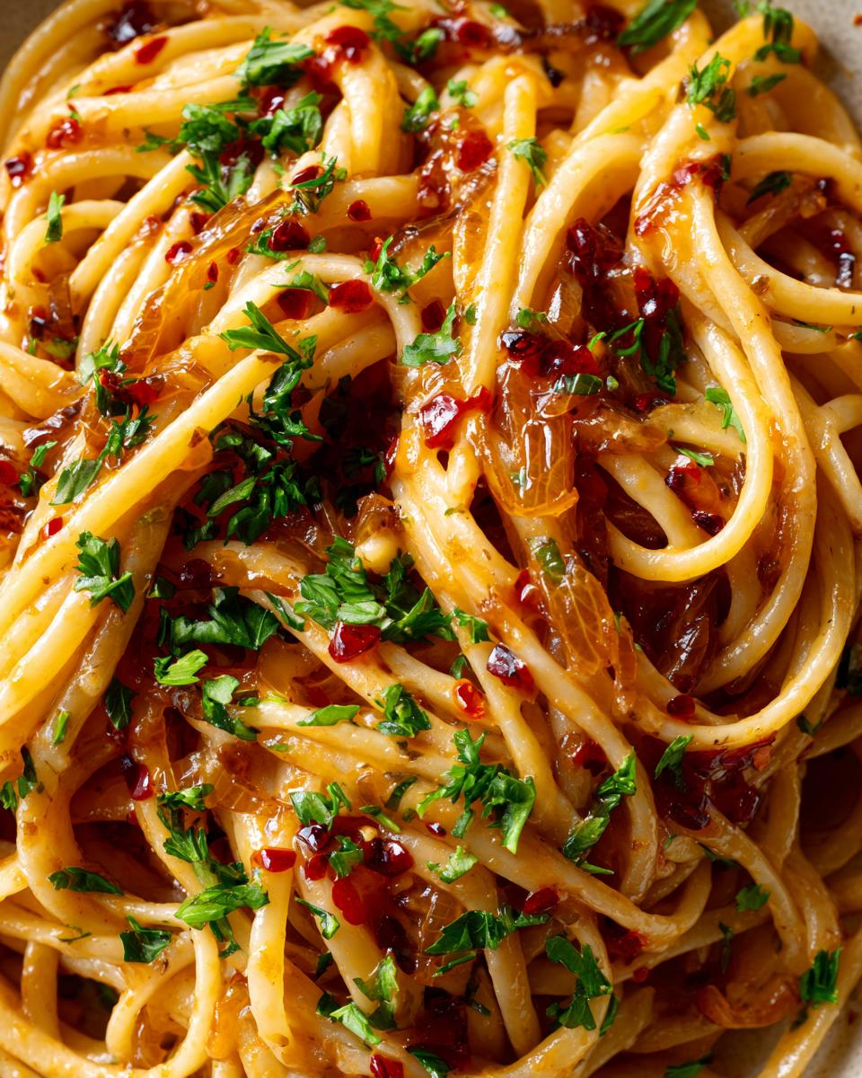 Close-up of thick spaghetti noodles tossed in a savory sauce with caramelized onion, chili flakes, and fresh parsley, showcasing the caramelized onion pasta.