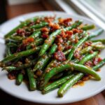 A close-up of glossy, bright green beans coated in a savory sauce and topped with crispy bacon bits.