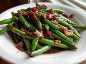 Close-up of bright green beans coated in a dark, sticky glaze and topped with crispy bacon pieces.