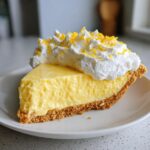 A thick slice of creamy, yellow lemon pie on a white plate, topped with whipped cream and lemon zest.