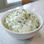 Close-up of a white bowl filled with creamy, chunky pickle dip topped with fresh dill.