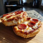 Two halves of freshly baked pepperoni pizza bagels with melted, gooey cheese resting on a wooden cutting board.