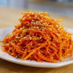 A close-up of a mound of bright orange raw carrot salad, shredded and topped with white sesame seeds.