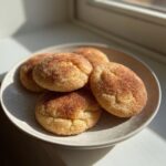 A stack of soft, round snickerdoodles generously coated in cinnamon sugar, resting on a light-colored plate.