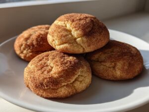 Four freshly baked, soft snickerdoodles generously coated in cinnamon sugar, stacked on a white plate.