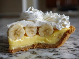Close-up of a perfect slice of banana cream pie showing the graham cracker crust, thick vanilla custard, banana slices, and piped whipped cream topping.