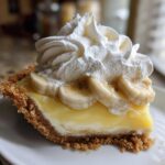A close-up slice of banana cream pie showing the graham cracker crust, creamy filling, banana slices, and tall swirl of whipped cream.
