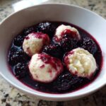 Four fluffy, light-colored Blackberry Dumplings served in a rich, dark purple sauce filled with fresh blackberries.