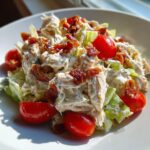 A close-up serving of creamy BLT chicken salad mixed with lettuce, topped with crispy bacon bits and halved cherry tomatoes.