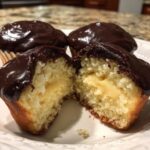 A boston cream pie cupcake cut in half showing the vanilla cake, pastry cream filling, and chocolate glaze.
