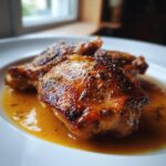 Close-up of two golden-brown, seasoned braised chicken thighs resting in a rich, savory sauce on a white plate.