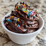 Close-up of rich, glossy brownie batter dip in a white bowl, topped with chocolate chips and colorful sprinkles.