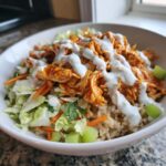 A close-up of a buffalo chicken bowl featuring shredded buffalo chicken, brown rice, lettuce, carrots, celery, and ranch dressing.