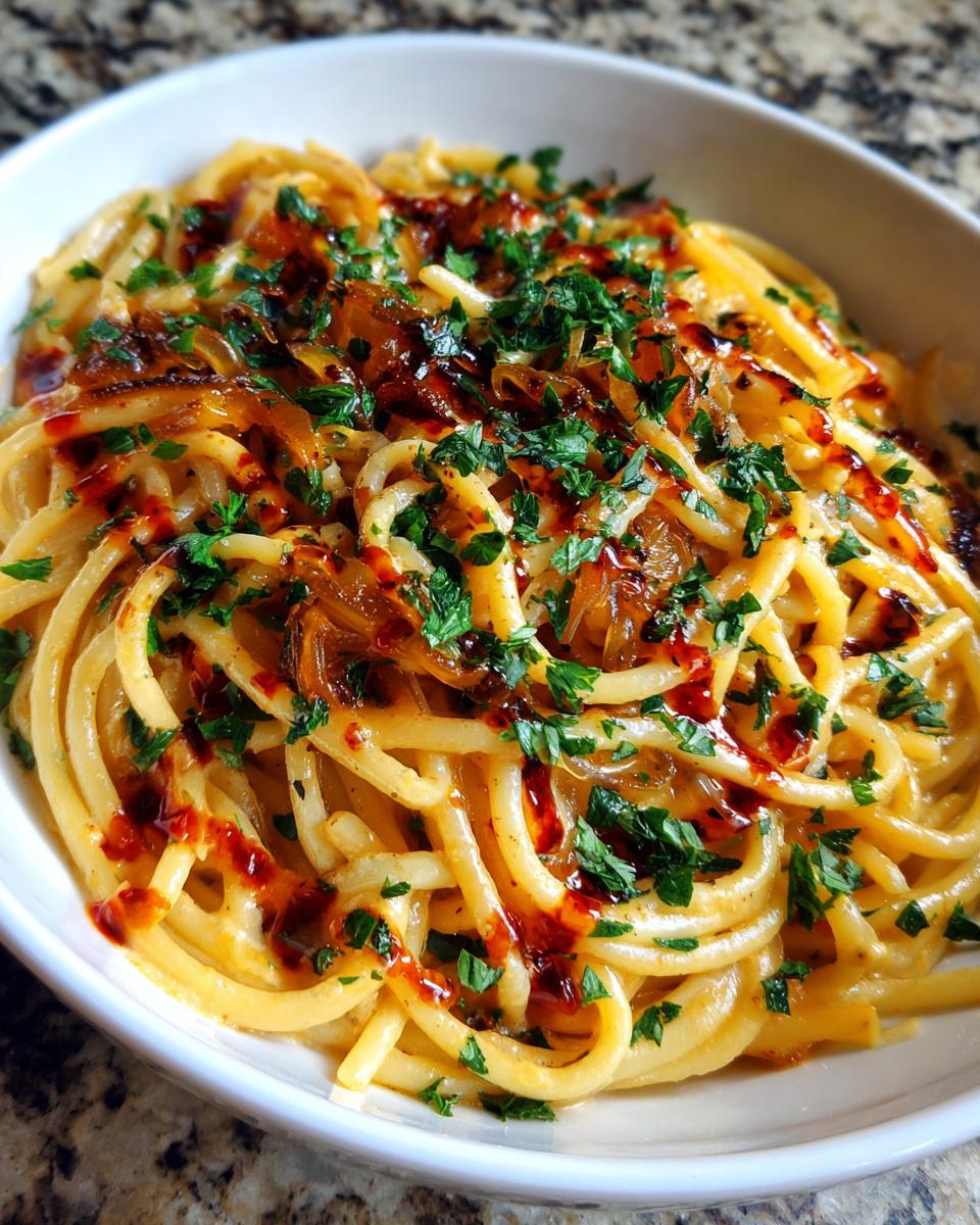 A close-up of creamy spaghetti tossed in sauce, topped with caramelized onion pasta, fresh parsley, and a drizzle of chili oil.