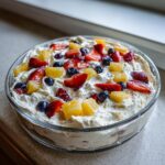 Close-up of a glass bowl filled with creamy cheesecake fruit salad topped with strawberries, blueberries, and pineapple chunks.