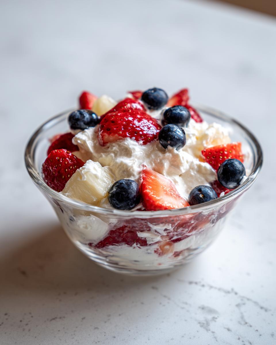 A glass bowl filled with cheesecake fruit salad featuring strawberries, blueberries, and pineapple mixed with cream.