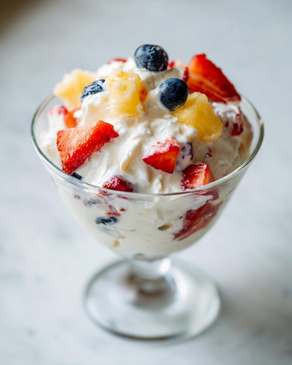 A close-up of a single serving of cheesecake fruit salad topped with strawberries, blueberries, and pineapple chunks.