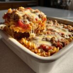 A serving spoon lifts a cheesy, saucy portion from a baking dish of pizza pasta bake.