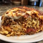 A close-up of a thick slice of pizza casserole featuring rotini pasta, seasoned ground meat, and a bubbly, browned mozzarella cheese topping.